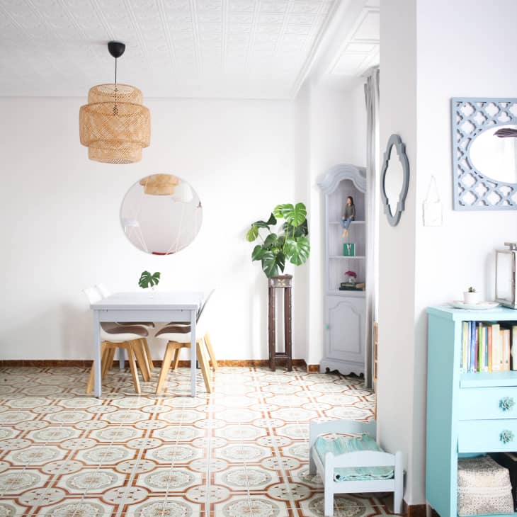 Dining room with white table, wooden chairs, round mirror, wicker pendant light, and plants on patterned tile floor.