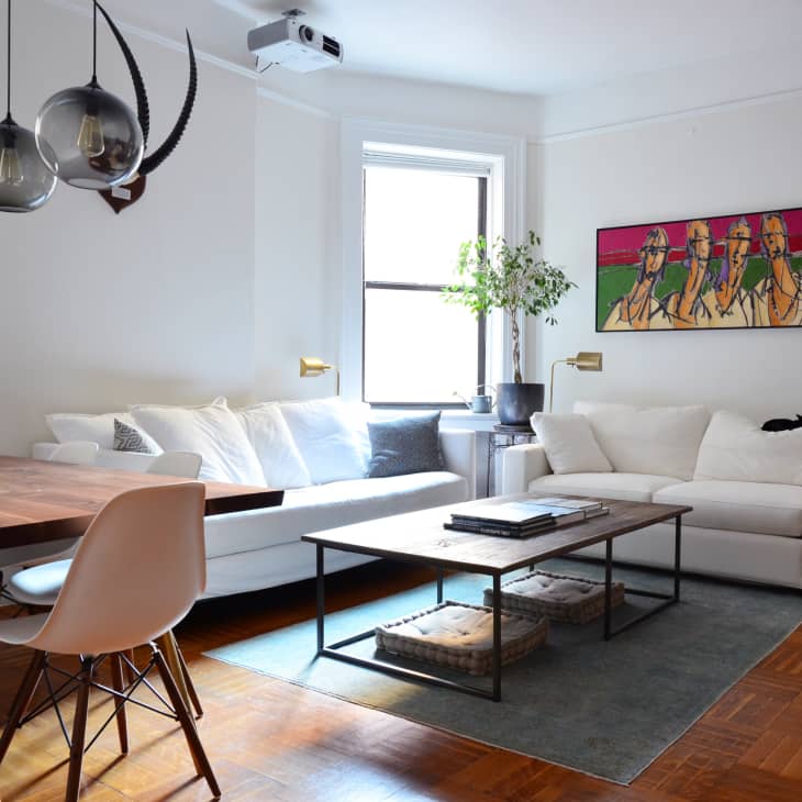 Modern living room with white sofas, abstract art, wooden table, pendant lights, and a potted plant by the window.