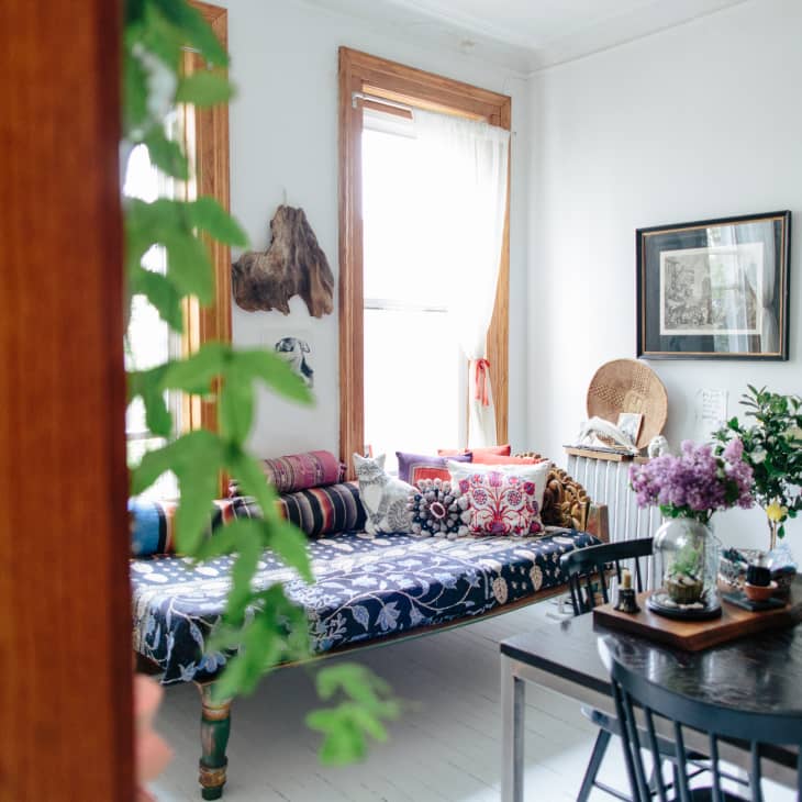 Cozy living room with a patterned daybed, colorful cushions, wooden dining table, and potted plants on a white mantel.