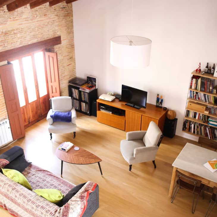 Living room with wooden floors, a gray sofa, armchairs, a TV on a wooden stand, bookshelves, and a dining table with chairs.