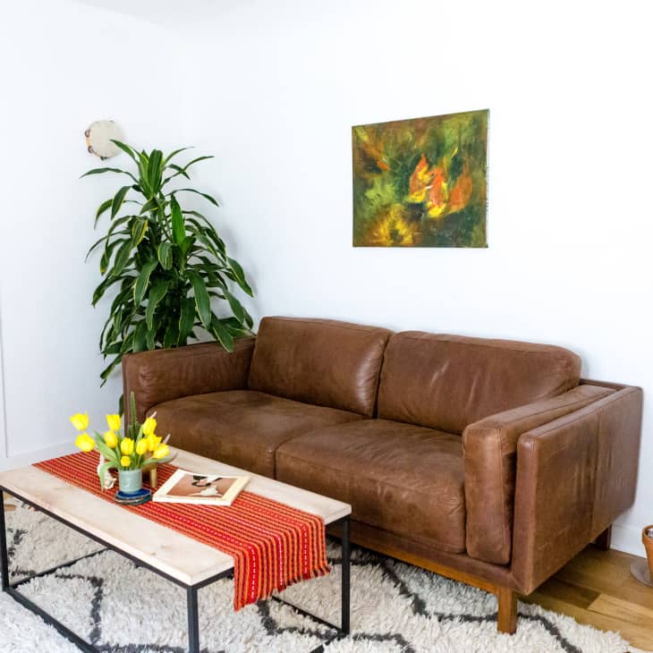 Brown leather sofa in living room with abstract art, potted plants, and a coffee table with yellow tulips and a red runner.