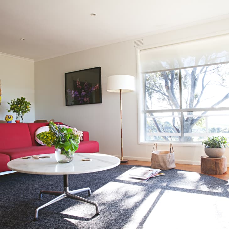 Bright living room with red sofa, white round table, floor lamp, and large window overlooking trees.