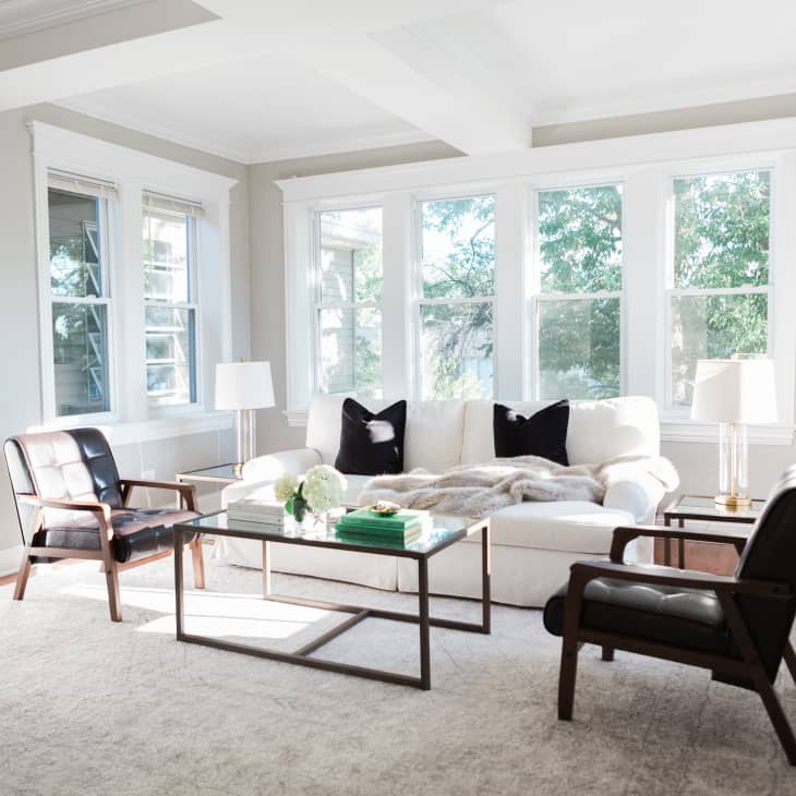 Bright living room with a white sofa, black pillows, glass coffee table, and two dark wooden chairs, featuring large windows.