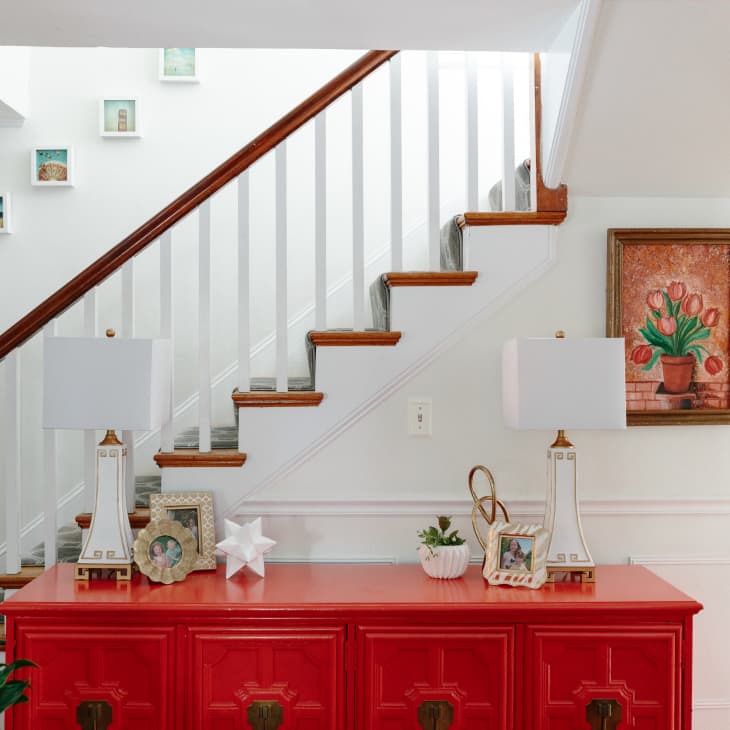 Red console table with lamps, decor, and framed photos beneath a staircase with a tulip painting on the wall.