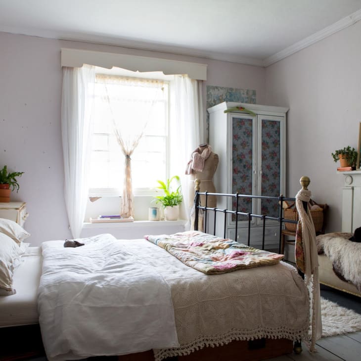 Cozy bedroom with iron bed, floral wardrobe, plants, and a cat on a fur-covered bench by the window.