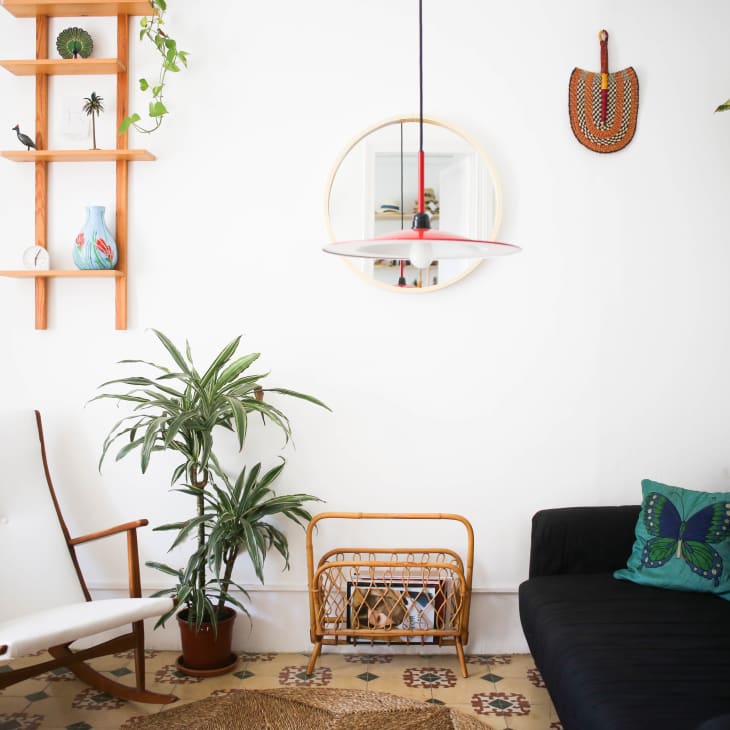 Living room with black sofa, butterfly pillow, white chair, wooden shelves, plants, and a round mirror.
