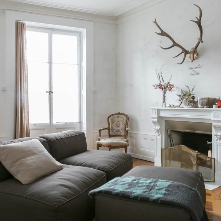 Cozy living room with gray sofa, white fireplace, antler decor, and a window with beige curtains.