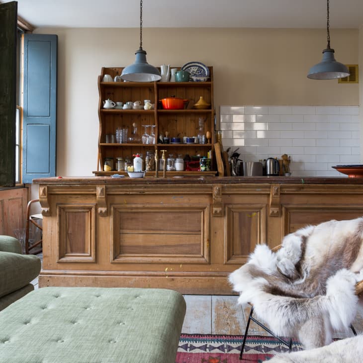 Cozy kitchen with wooden island, open shelves, green sofa, fur-covered chair, and large windows with blue shutters.