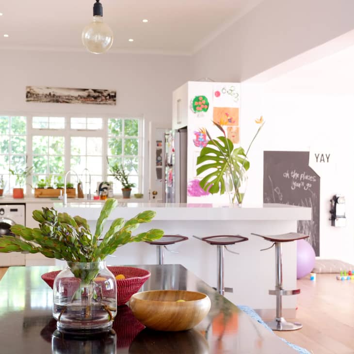 Bright kitchen with white cabinets, island with bar stools, large plant, and colorful artwork on the walls.