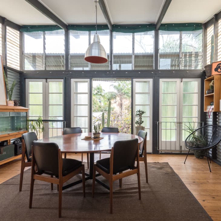 Dining room with round wooden table, six chairs, large fish tank, bookshelves, and plants, surrounded by glass doors.
