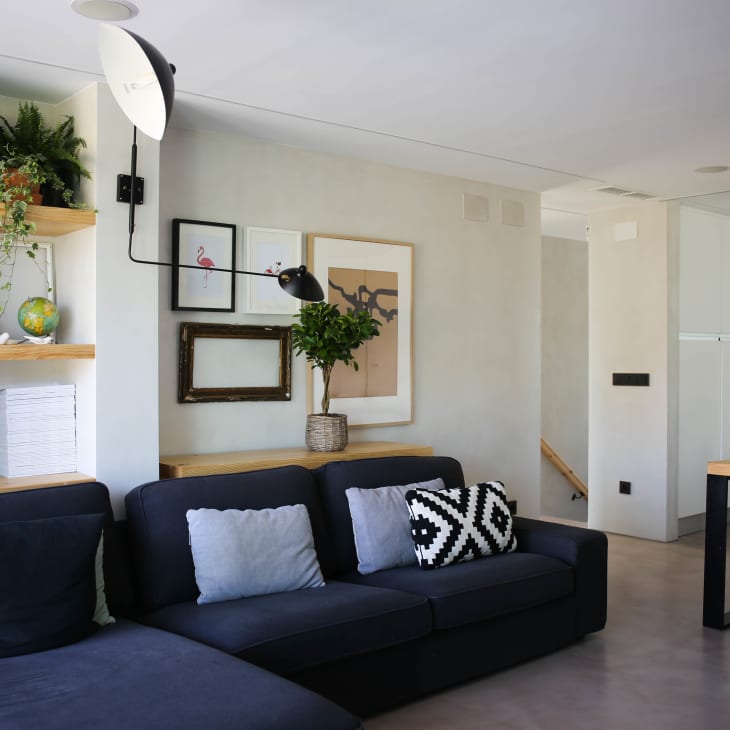 Modern living room with dark sofa, geometric cushions, wall art, and wooden shelves with plants and books.