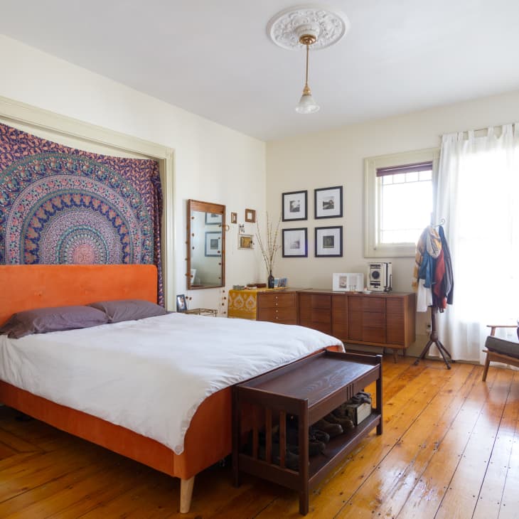 Bedroom with orange headboard, tapestry, wooden dresser, and desk by a window with sheer curtains.