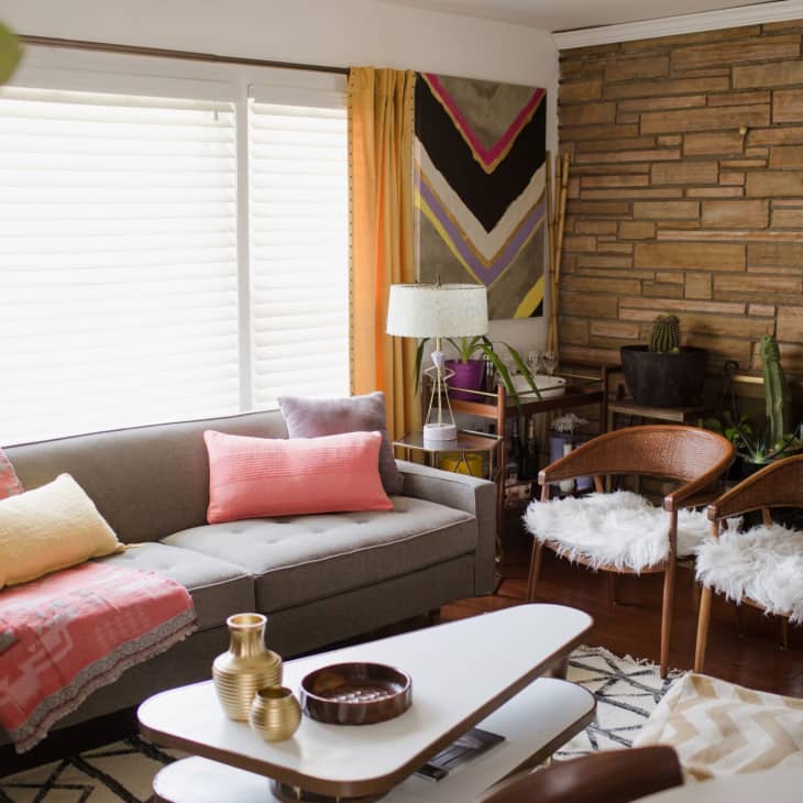Mid-century modern living room with gray sofa, colorful pillows, wooden chairs, and potted plants against a stone wall.