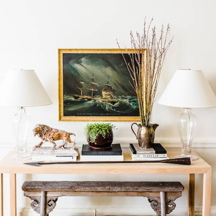 Console table with two lamps, a ship painting, books, a lion sculpture, and a vase with branches.