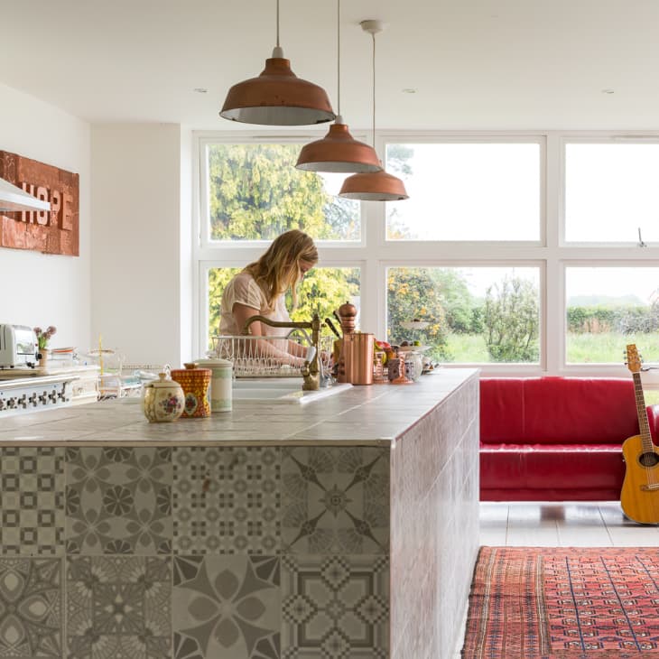 Kitchen with patterned island, woman preparing food, red sofa, guitar, and large windows overlooking garden.
