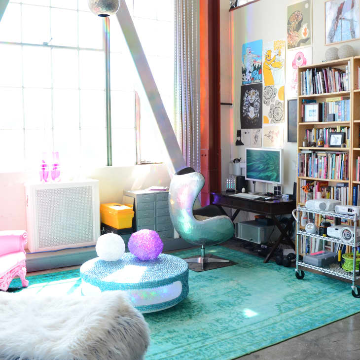Colorful living room with pink armchair, fluffy white chair, teal rug, bookshelves, and a modern desk setup.
