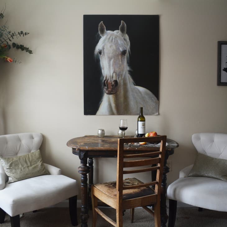 Cozy dining nook with a round wooden table, two white chairs, wine bottle, and horse painting on the wall.