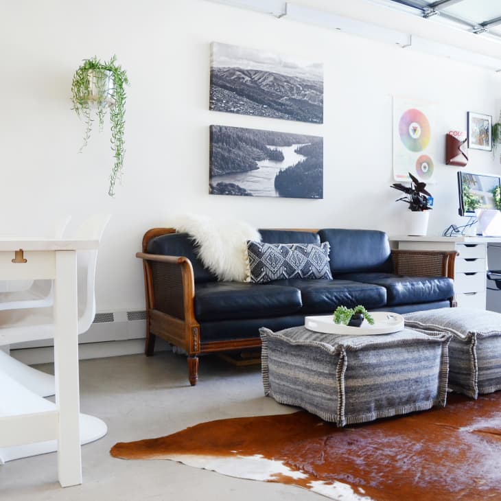 Modern living room with black leather sofa, white desk, computer, and wall art, featuring plants and a cowhide rug.