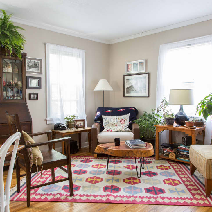 Cozy living room with patterned rug, wooden furniture, plants, and framed photos on beige walls.