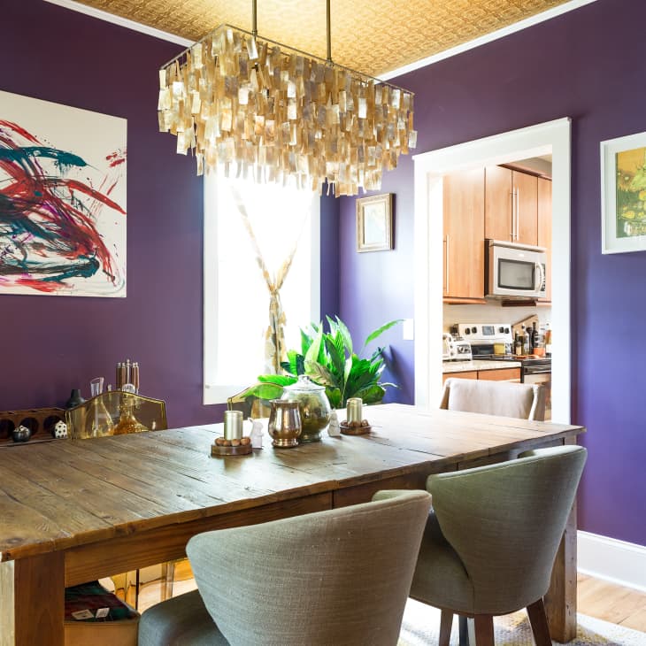 Dining room with purple walls, abstract art, wooden table, gray chairs, and a modern chandelier. Kitchen visible through doorway.