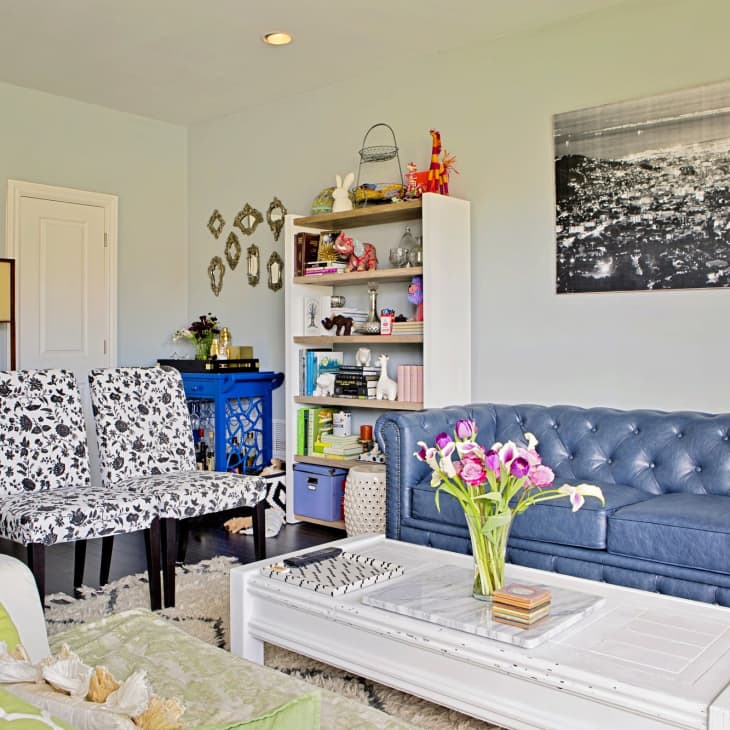 Living room with blue tufted sofa, floral chairs, white coffee table, and a bookshelf. Black and white cityscape on wall.