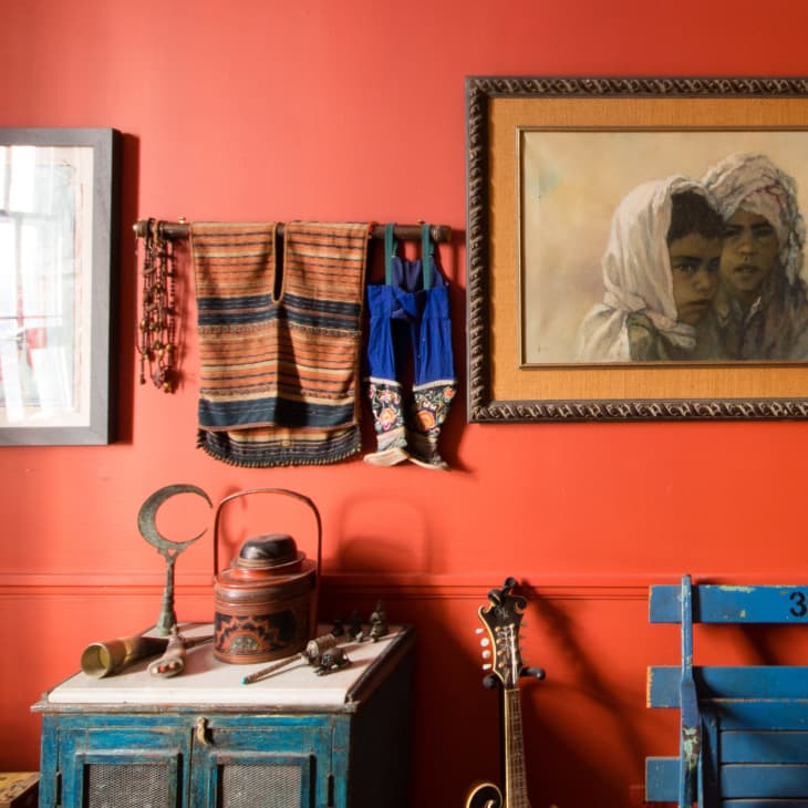 Eclectic room with red walls, framed art, textiles, a blue chair, and a guitar leaning against a vintage cabinet.