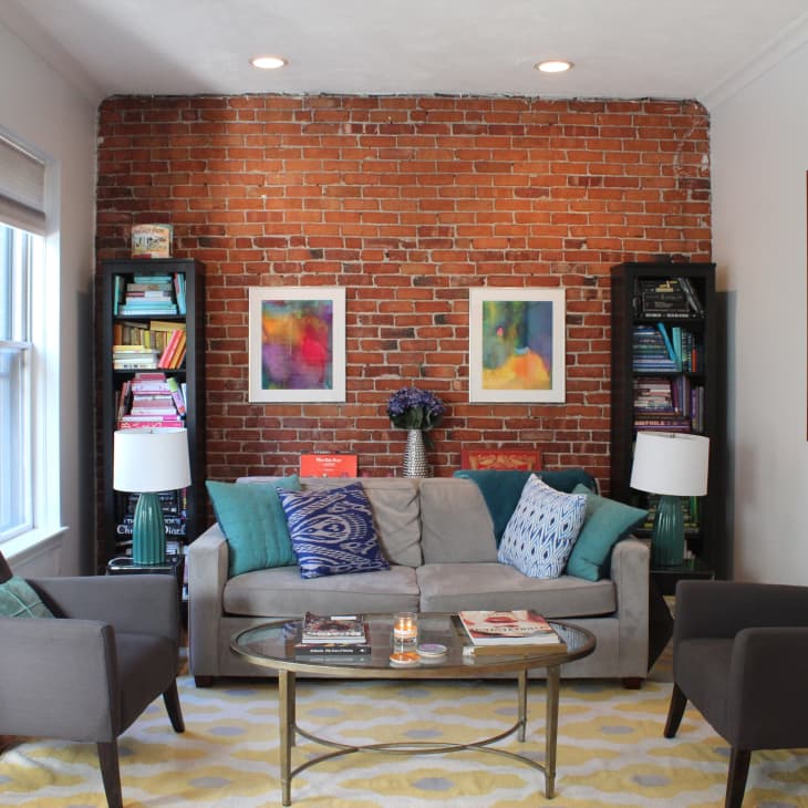 Cozy living room with exposed brick wall, gray sofa, colorful cushions, two armchairs, bookshelves, and abstract art.