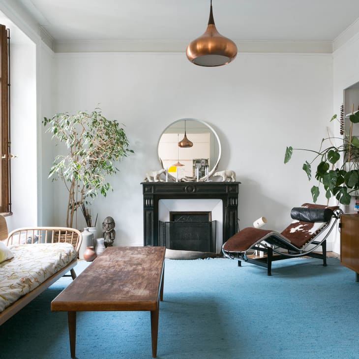 Living room with a wooden sofa, blue rug, black fireplace, large plants, and a copper pendant light.