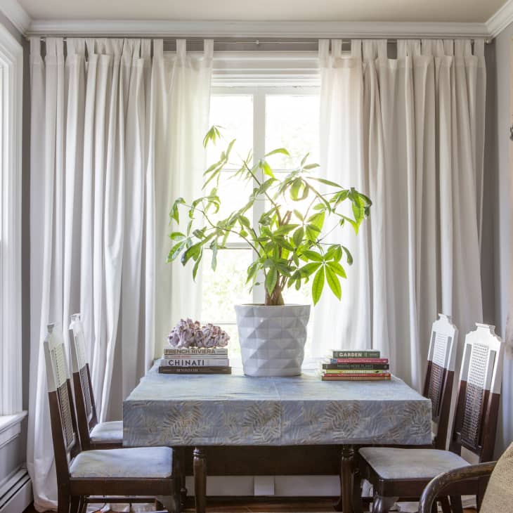 Dining room with a table, potted plant, books, and four chairs, framed by white curtains and a large wall art piece.