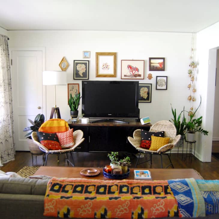 Living room with a TV, colorful cushions on chairs, wall art, plants, and a patterned throw on a gray sofa.