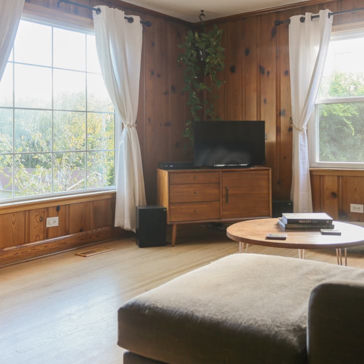 Living room with wood paneling, large windows, a TV on a wooden stand, a round coffee table, and a beige sofa.