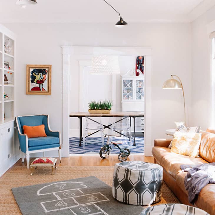 Living room with tan leather sofa, blue armchair, hopscotch rug, and a tricycle near a dining area with a table.