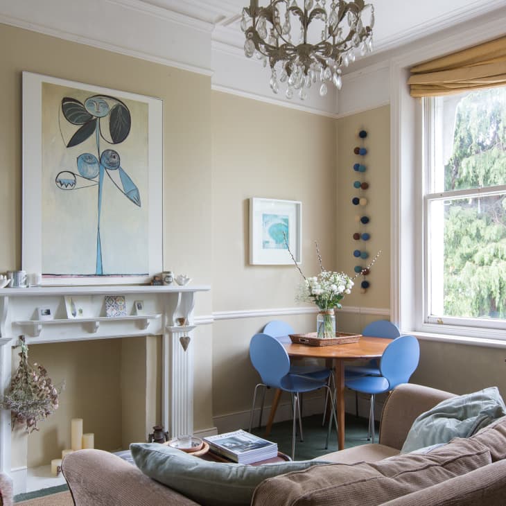 Living room with beige walls, blue chairs, round table, large artwork, chandelier, and a cat on the windowsill.