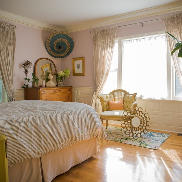 Sunlit bedroom with a large bed, wooden dresser, decorative mirror, and a chair by the window with sheer curtains.