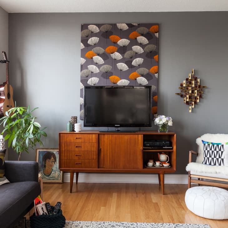 Mid-century modern living room with a TV on a wooden console, patterned wall art, guitar, and a cozy chair with a pouf.