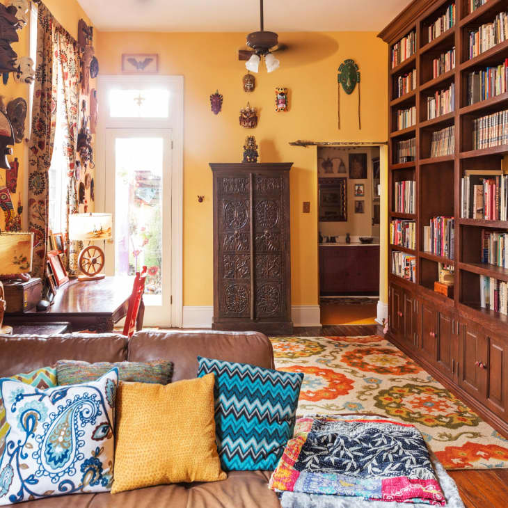 Living room with yellow walls, tribal masks, a brown leather sofa, colorful pillows, a large bookshelf, and a patterned rug.