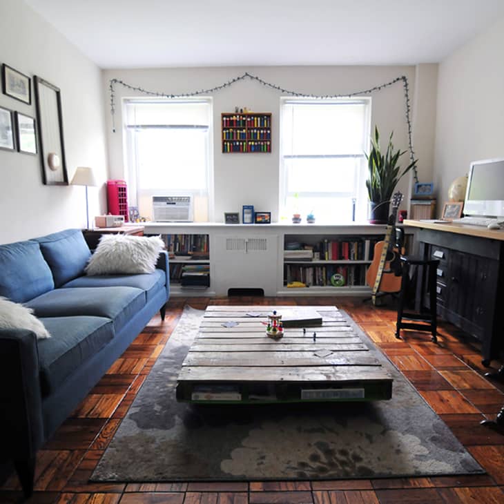 Living room with blue sofa, pallet coffee table, wall art, and a desk with a computer, books, and a guitar by the window.