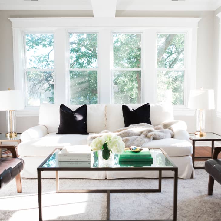 Bright living room with white sofa, black pillows, glass coffee table, and two leather chairs, surrounded by large windows.