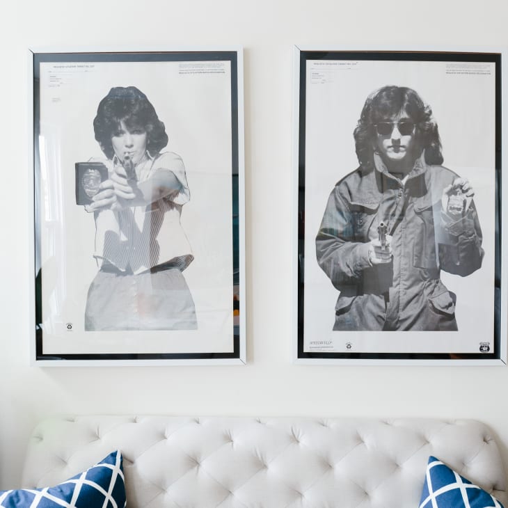 Two framed black-and-white posters of people holding guns above a white tufted sofa with blue patterned pillows.
