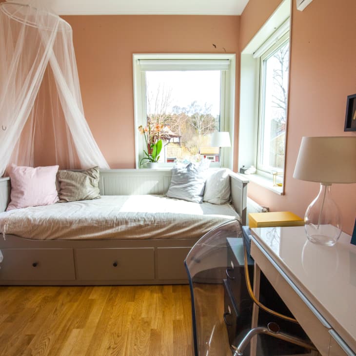Cozy bedroom with pink walls, daybed with canopy, desk, clear chair, and table lamp by a window.