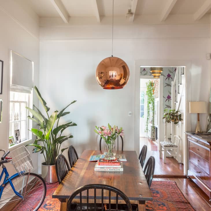 Dining room with wooden table, black chairs, copper pendant light, large plant, blue bicycle, and framed photos on wall.