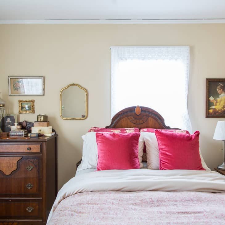Bedroom with wooden dresser, ornate headboard, pink pillows, framed art, and a table lamp on a nightstand.