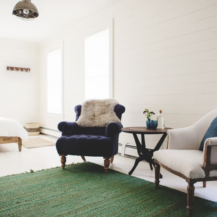 Bright bedroom with a navy armchair, white armchair, round table, and green rug.