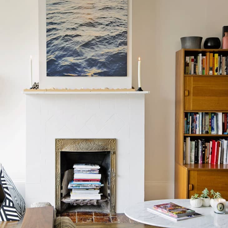 Living room with a white fireplace, ocean artwork, wooden bookshelf, and a round marble coffee table with books and plants.