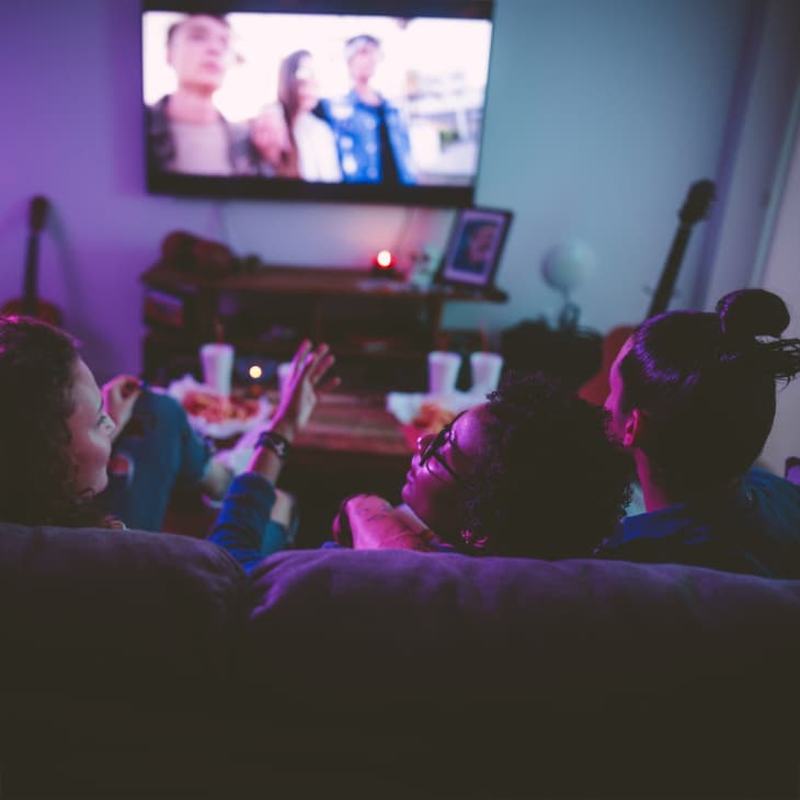 Young multi-ethnic hipster friends relaxing on living room sofa and watching film on TV