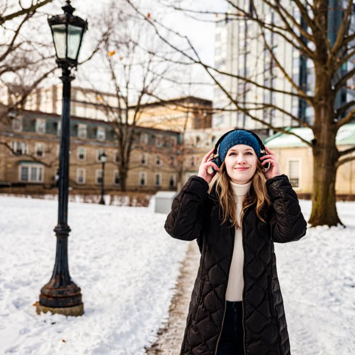 Female university student with headphones, listening to favourite tunes. Exterior of city park with historic buildings in the background.