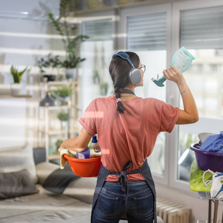 Woman with wireless headphones holding a basket with cleaning products and singing in living room at home.