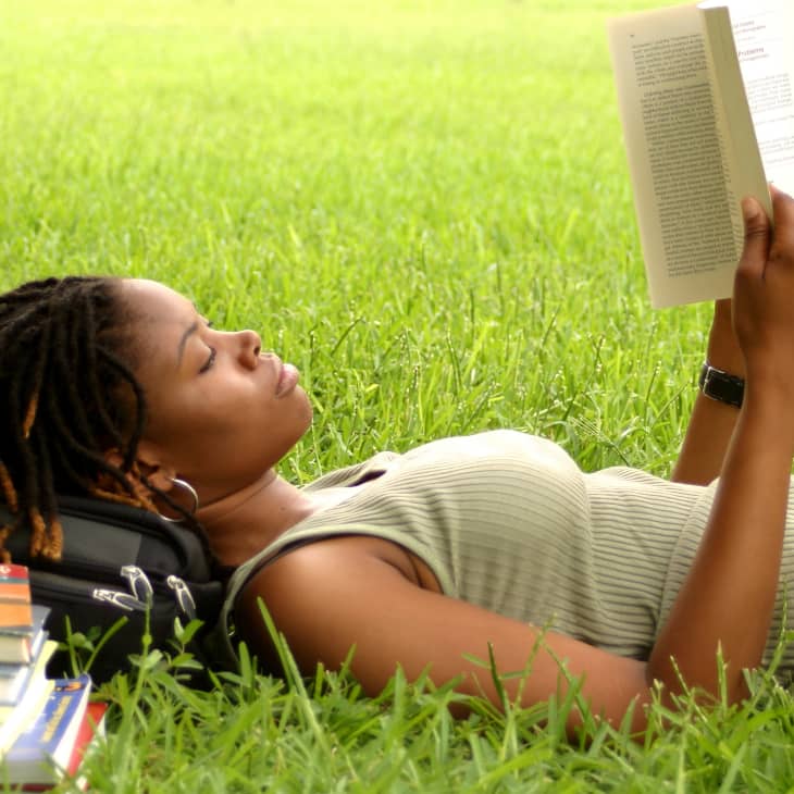 Young woman (college student) lying on grass, reading book, side view