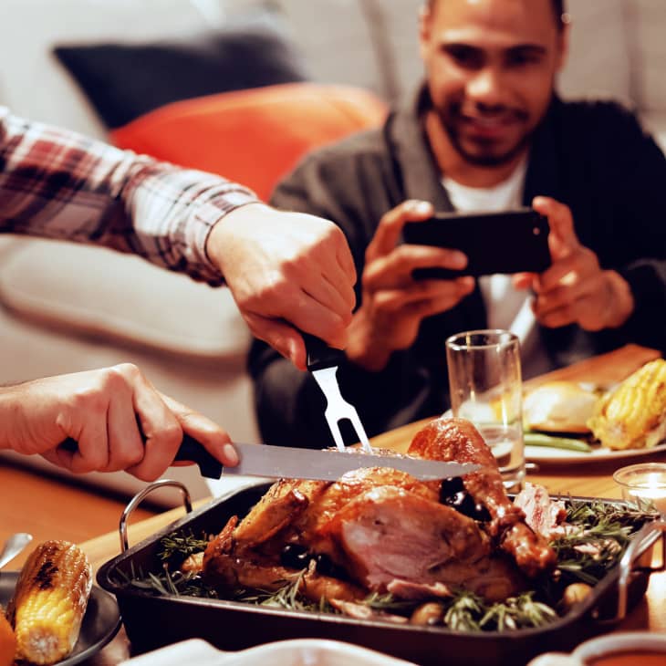 Front view of a young mixed race man sitting at a table for Thanksgiving dinner at home with friends carving the turkey, a young mixed race male friend taking a photo with his smartphone in the