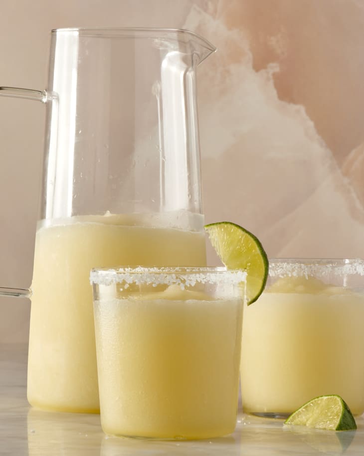 photo of a pitcher of frozen margarita cocktail on a marble surface with two salt rimmed glasses with lime wedge garnishes